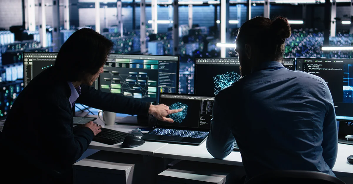 Two people monitoring system dashboards and security alerts at a workstation, representing cybersecurity operations and organisational risk visibility.